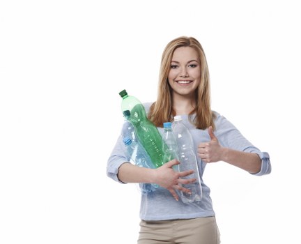A young woman with shoulder-length red hair and fair skin is standing against a plain white background. She is smiling and giving a thumbs-up gesture with her right hand. She is wearing a light blue long-sleeved shirt rolled up at the sleeves and beige trousers. In her left arm, she is holding several clear plastic bottles of various sizes, some with green or blue caps, and one green plastic bottle appears partially crushed. The bottles are associated with household waste commonly encountered in rubbish removal services, and her cheerful expression suggests satisfaction with waste collection or recycling efforts. This image is relevant for a rubbish removal service operating in Harpenden or nearby areas, supporting clean and environmentally responsible waste disposal practices.