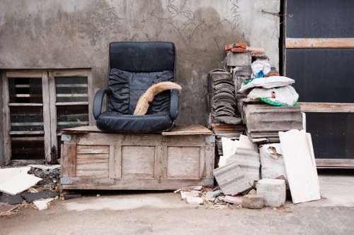 A black office chair with a cushioned seat and backrest, positioned on a wooden crate or pallet, situated outdoors against a weathered, beige concrete wall with visible stains and marks. To the right of the chair, there is a pile of construction debris including several stacked and leaning gray concrete blocks, some with rough, textured surfaces, and a few white panels or slabs among the rubble. The ground appears to be a concrete or paved surface with small scattered broken pieces of debris around the base of the pile. In the background, there are part of a vent or grille inset into the wall and a black door with a wooden horizontal reinforcement which suggests an industrial or back alley setting, typical of rubbish clearance scenes in towns like Harpenden. The overall scene indicates a collection of discarded office furniture and building waste awaiting removal by a professional rubbish clearance service such as House Clearance Harpenden.