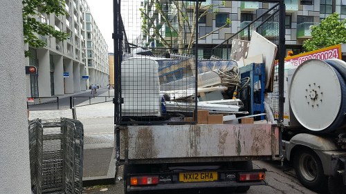 A flatbed truck parked on a city street in Harpenden, loaded with various waste materials including large white and beige panels, metal scraps, and wooden items, all secured with metal fencing to contain the rubbish during transportation. The truck's rear is directed towards the viewer, displaying the license plate 'NX12 GHA,' and is positioned adjacent to a modern building with glass balconies and landscaping features. The surrounding environment includes a paved sidewalk and an intersection visible in the background, with a traffic light and street signs indicating a busy urban area. This scene exemplifies the type of rubbish removal service provided by House Clearance Harpenden, capturing the process of collection and transportation of household or commercial waste in the local area.
