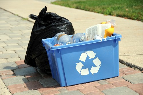 An outdoor scene featuring a blue plastic recycling bin with a white recycling symbol on the front, positioned on a paved pathway near a grassy area. The bin is filled with transparent plastic bottles, some with visible caps, and a few paper and cardboard items. Next to the bin, there is a black plastic rubbish bag, tightly tied at the top, leaning against the bin. In the background, a concrete sidewalk and a grassy verge are visible, suggesting a residential or suburban setting. The lighting indicates a bright day, emphasizing the contrast between the bin's bright blue colour and the darker rubbish bag, typical of rubbish removal tasks undertaken by companies like House Clearance Harpenden, serving the Harpenden area.