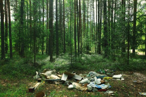 A pile of various household rubbish and discarded debris lies on the ground in a forested area, with tall trees and dense green foliage surrounding the scene. The waste includes cardboard boxes, plastic bags, paper remnants, and other mixed trash, scattered across the uneven forest floor, which is covered with leaves and small plants. The rubbish appears haphazardly dumped, with some items overlapping and partially crumpled. Sunlight filters through the canopy of trees, illuminating the area with natural light. This scene exemplifies improper disposal of waste in an environment that typically remains untouched. House Clearance Harpenden's rubbish removal services are designed to prevent such littering and ensure proper disposal, especially in urban or semi-rural areas near Harpenden, Hertfordshire. The image highlights an environment affected by illegal dumping, emphasizing the importance of professional rubbish clearance to maintain cleanliness and protect local natural surroundings.
