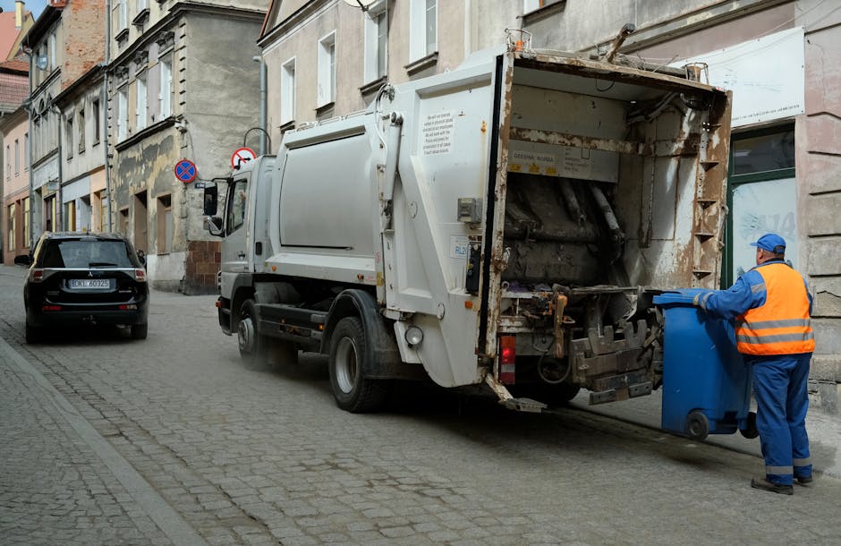 A waste collection truck is parked on a narrow urban street, positioned close to the curb with its rear compartment open, revealing an empty, rusted, and weathered metal interior designed for rubbish
