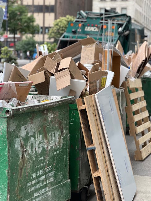 A waste collection truck is parked on a narrow urban street, positioned close to the curb with its rear compartment open, revealing an empty, rusted, and weathered metal interior designed for rubbish