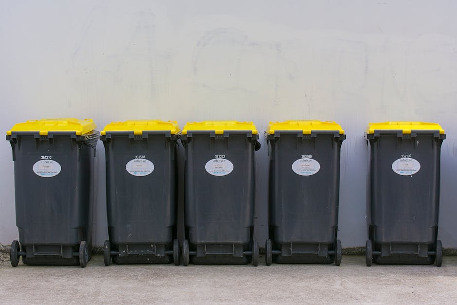 Close-up of a tightly tied black plastic garbage bag with a glossy surface, placed against a plain, light-colored wall. The bag appears full, with creases and folds indicating it contains waste materi
