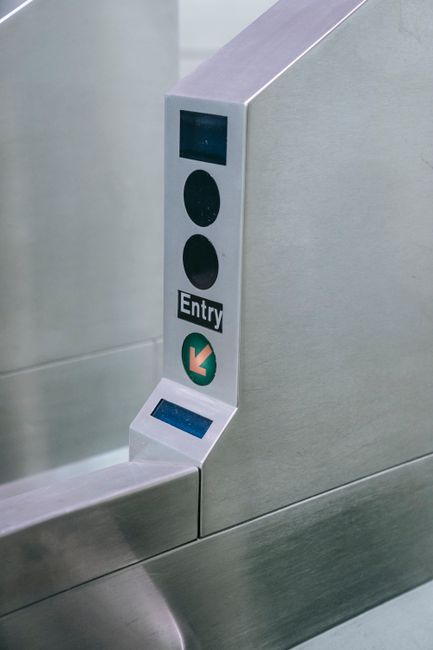 A close-up view of a modern elevator control panel mounted on a light grey metallic wall. The panel includes a small rectangular digital display screen at the top, two circular black buttons, with the