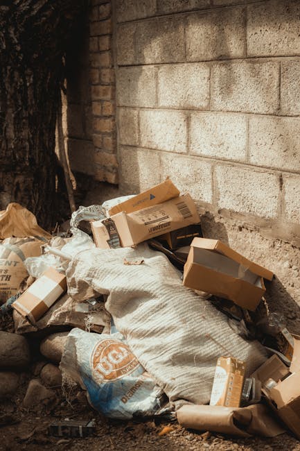 A scene of outdoor debris featuring a mix of household waste and large discarded items. In the foreground, there is a small, red plastic toy car with a black steering wheel, scratches, and worn paint,